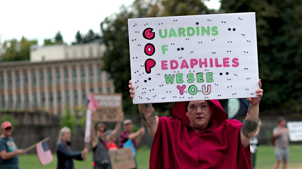 Olympia Washington, Photo of person holding protest sign, Guardins of Pedaphiles, We See You, numerous sets of Googly Eyes on sign, sign being held by person wearing red robe with red hood