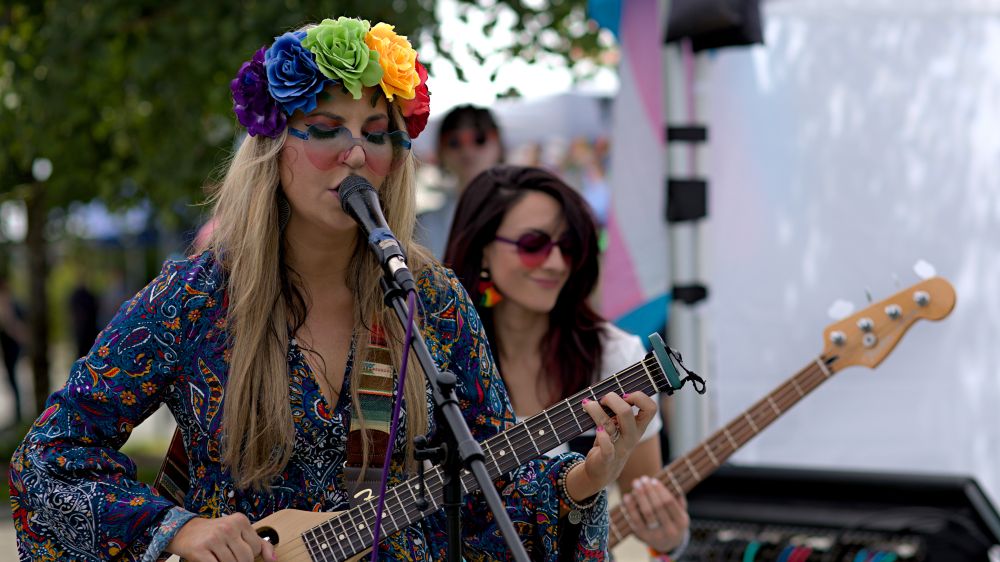 Photo of musician at microphone signing, heart sunglasses, playing guitar, multi pride color roses on head, purple blue, green yellow, red - another guitarist in background, trans flag seen