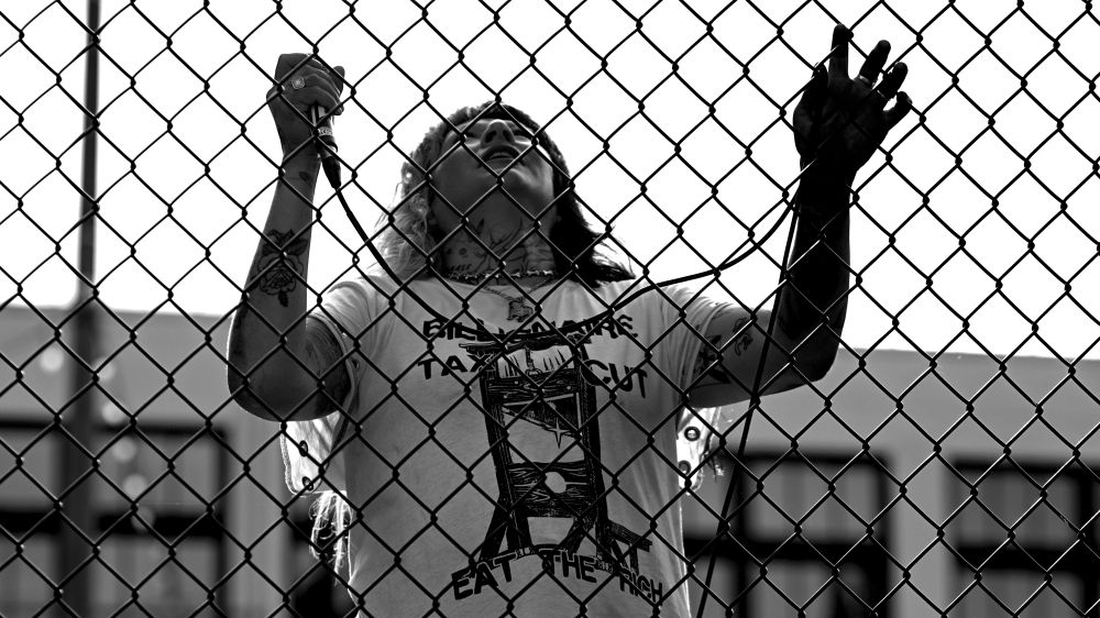 Monochrome photo of musician with microphone holding onto fence at Cal Anderson Park, looking up. Musician wearing t-shirt Billionaire Tax Cut, picture of guillotine, eat the rich.