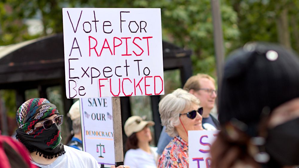 Person holding protest sign, Vote for a rapist expect to be fucked, protesters seen in background
