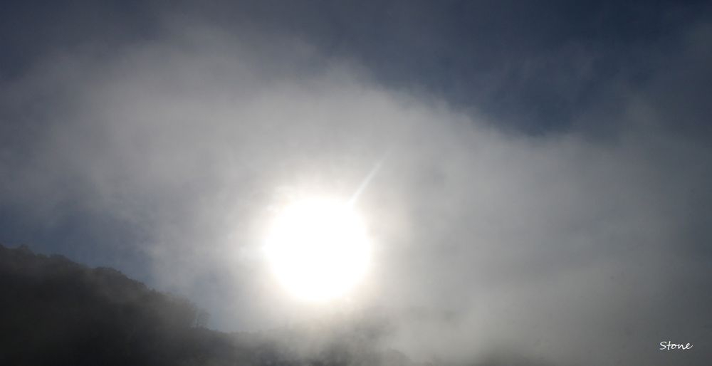 the sun shining through fog. Can see a bit of a hill on the bottom left. & sorta see the blue sky through the fog.