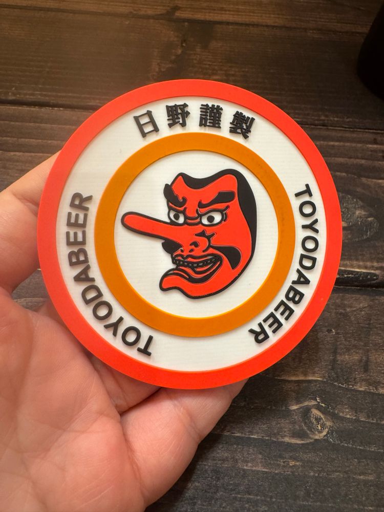 コースターのアップ。天狗の顔と「日野謹製」「TOYODA BEER」の字。