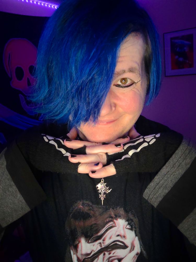 Local Twitter/YouTube/TikTok/Bluesky sensation Super Nerd Daniel, posing for a photo in front of their cell phone's camera with a slight smile on their face and their chin resting on top of the interlocked fingers of his hands.

Daniel is a white nonbinary person with dyed blue hair and hazel-brown eyes, wearing a black t-shirt of WWE professional wrestler Tatum Paxley (depicting two Tatums splitting apart from each other in the style of the Junji Ito horror manga "Tomie") over a long-sleeved striped black-and-grey shirt, black fingerless gloves with white skeleton hand patterns on them, black fingernail polish, black eyeliner with small "wings" protruding from the edges, and a small silver necklace modeled after that of Squall Leonhart from the video game Final Fantasy VIII.