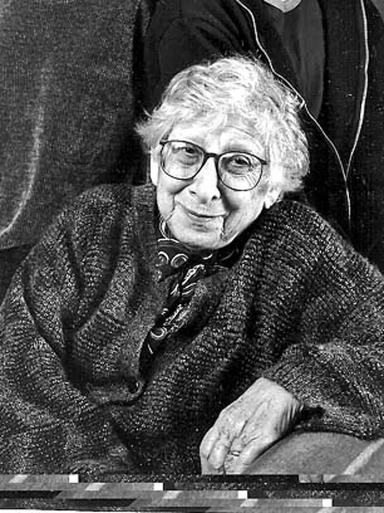 A black and white image of a woman in her 80s with large glasses and a rye smile, seated and leaning on the arm of a chair.