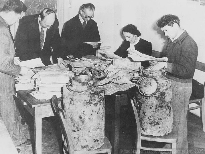 Schwarz-Weiß-Fotografie von fünf Personen - vier Männern und einer Frau - die um einen Tisch stehen bzw. sitzen, auf dem zahlreiche Dokumente ausgebreitet sind. Interessiert lese sie in den Dokumente. Vor dem Tisch, auf zwei Stühlen, stehen zwei Milchkannen.