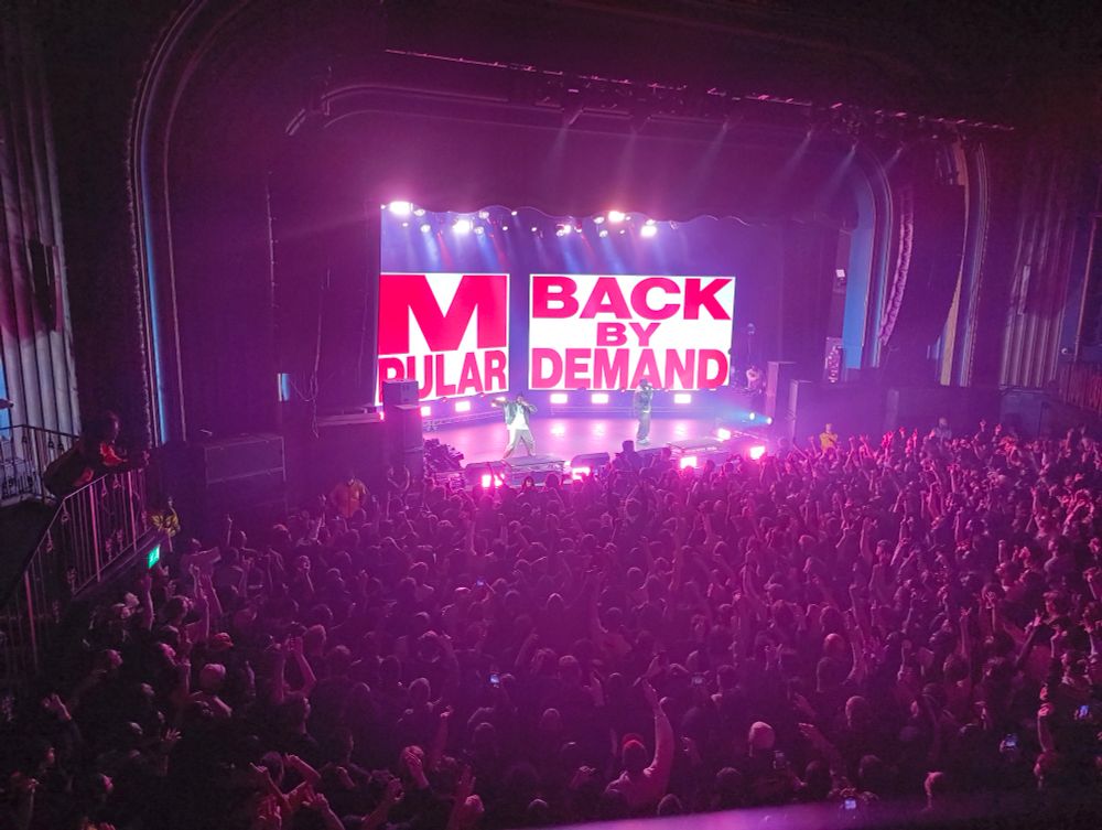 A view from the balcony of the Kentish Town Forum looking down on a large crowd with Clipse on stage, the background visuals show large red lettering saying 'Back by popular demand'