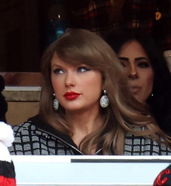 taylor swift looking stunning wearing a black and grey plaid jacket, big white dangly earrings, her classic red lip, and her hair down with a side bang while sitting down at a chiefs game