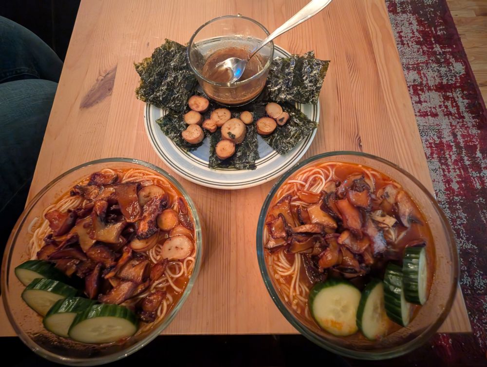 Two bowls of ramen with mushrooms and cucumbers on top. A plate with seaweed, fried mushroom rounds and a ramekin of garlic butter sauce.