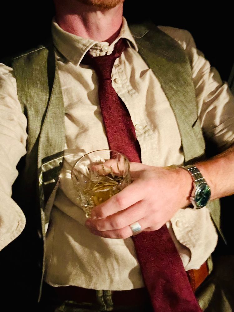 Mein Oberkörper mit beigen Hemd, grüner Weste und weinroter Paisley Krawatte. In meiner Hand ein Glas Scotch.