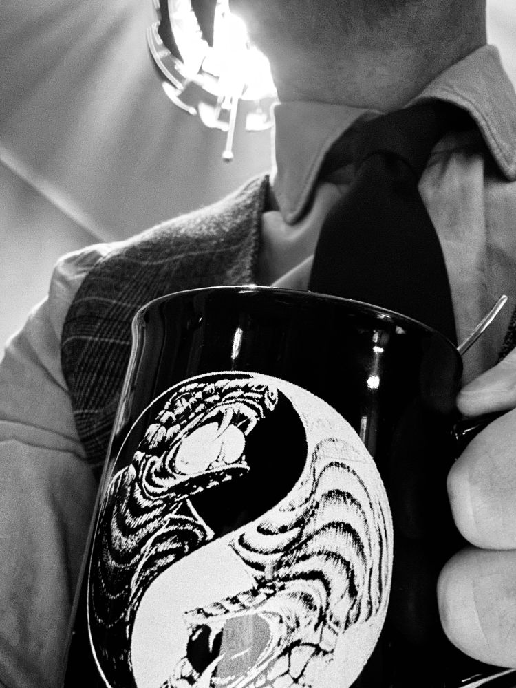Ich sitze mit Hemd, Krawatte und Weste mit einer Tasse Kaffee in der Hand. Man sieht mich bis zum Hals, über mir die leuchtende Lampe.