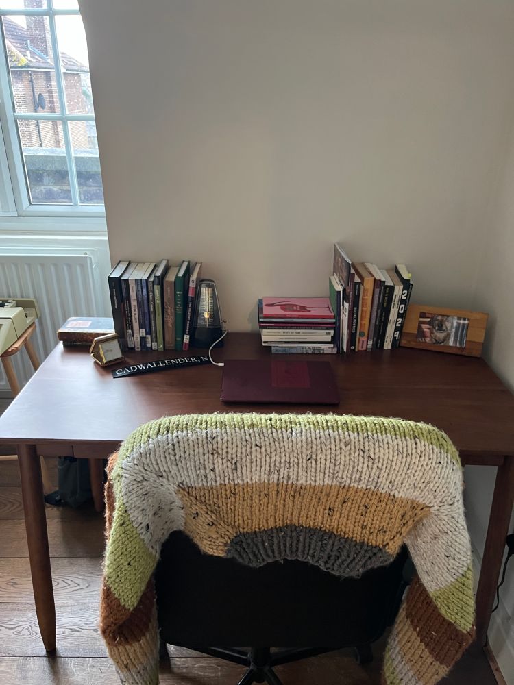 A wooden desk with books, laptop, a chair with a sweater on it, by a sunlit window.
