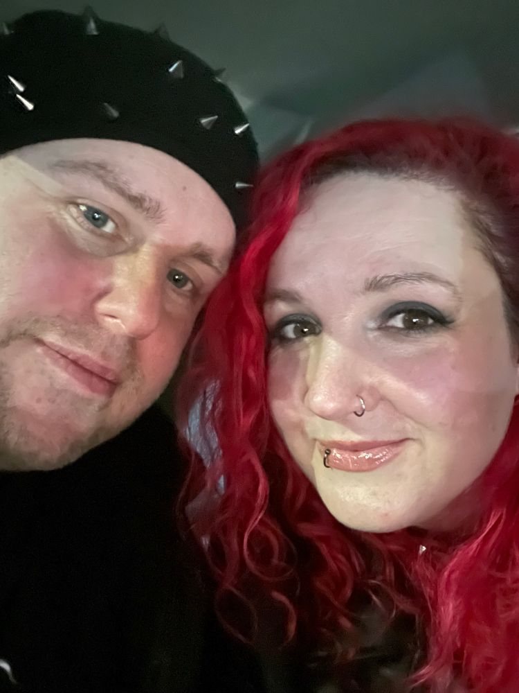 A selfie of Jenny and her husband, Dave. 
Jenny has long, bright red,  curly hair and has dark smokey eye make up. 
Dave is wearing a black beanie with spikes. Both are smiling at the camera 