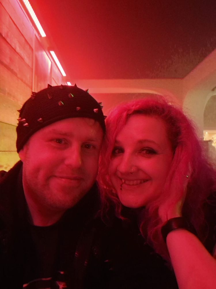 A selfie of Jenny and her husband, Dave. Taken in a club so the lighting is very red. Dave wears a black beanie with spikes. Jenny has long red hair. Both are smiling at the camera 