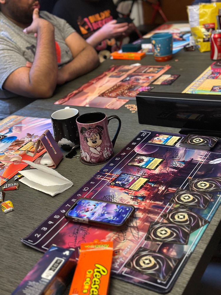 Long table with Lorcana playmats and cards. Coffee cups and snacks are visible as well. 