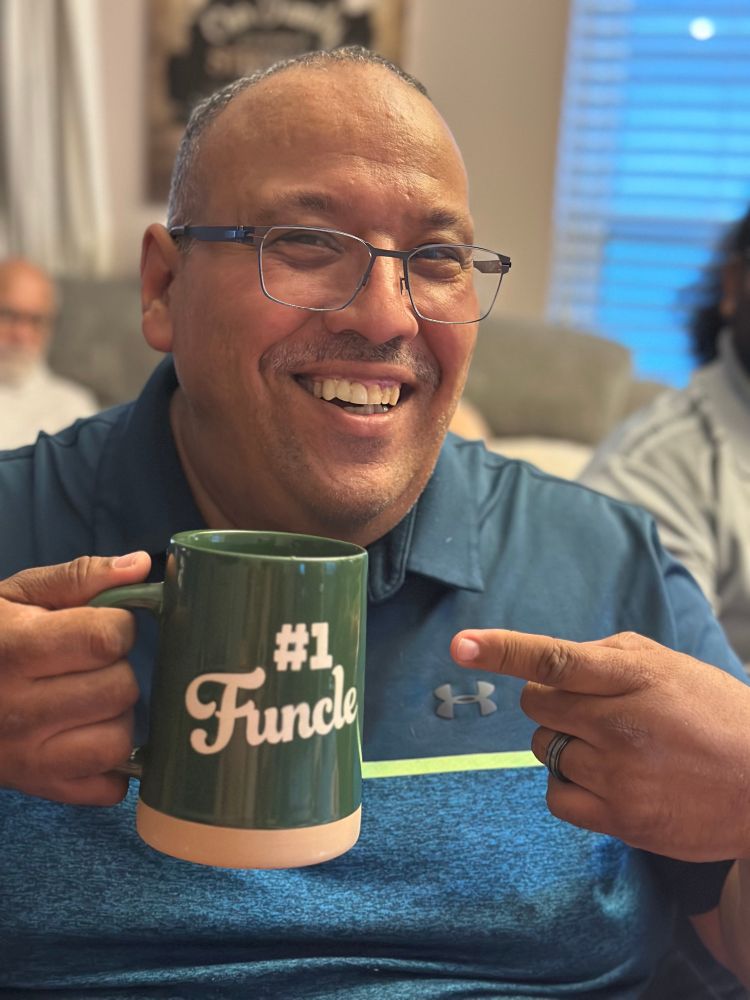 A very happy man is smiling while pointing at the large coffee mug in his hand that says Number One Funcle. 