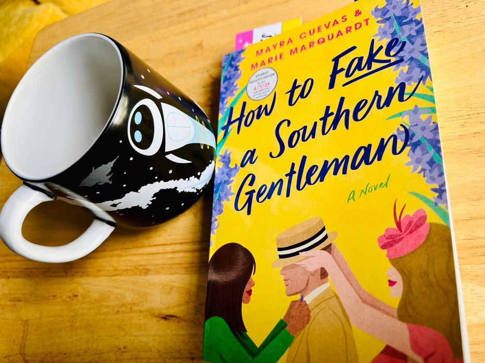 A coffee mug with Eve from Wall-E sits next to an advanced reader copy with an illustrated cover of HOW TO FAKE A SOUTHERN GENTLEMAN by Mayra Cuevas and Marie Marquardt.