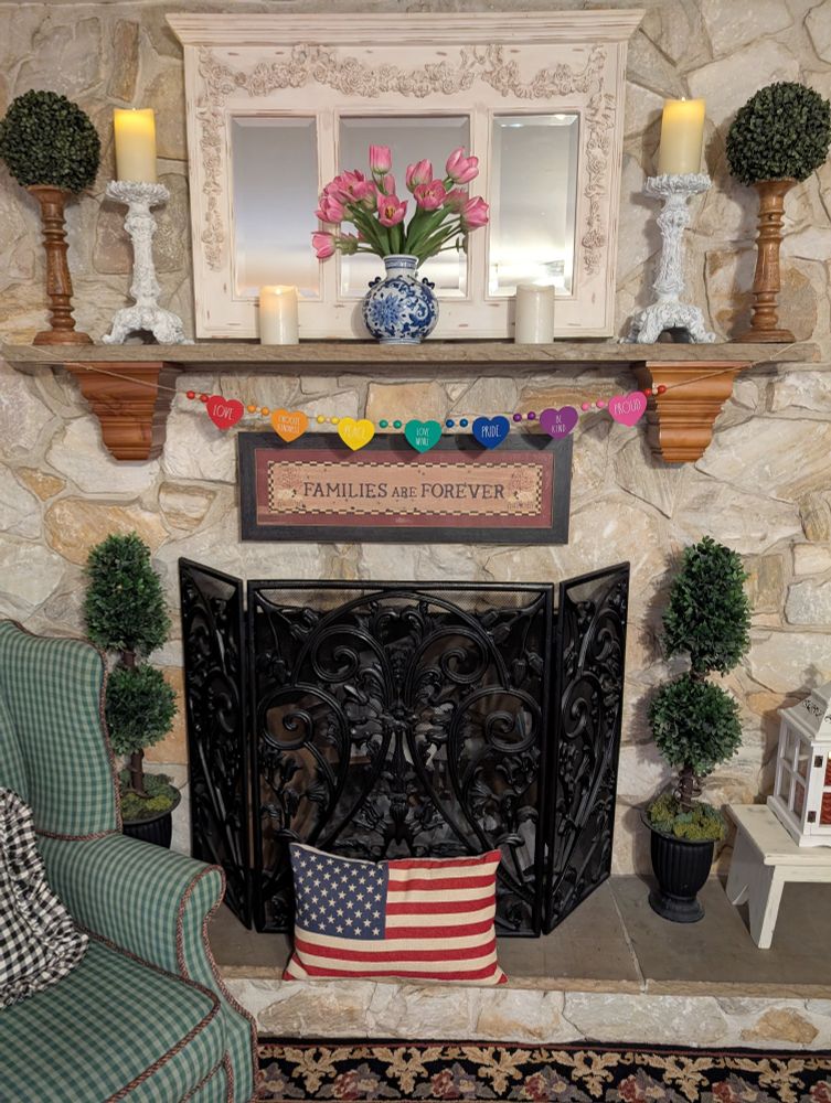 Picture of a fireplace with a farm house sign saying "Families Are Forever", American flag pillow, and wooden pride 7 hearts on a string in each color of the rainbow with beautiful sayings and words. Red: Love, Orange: Choose Kindness, Yellow: Peace, Green: Love Wins, Blue: Pride, Purple: Be Kind, Pink: Proud. 