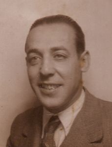 Foto de Emilio. Primer plano de él sonriendo cuando tenía, quizás unos 30 años. Lleva camisa, corbata y traje y está muy repeinado hacia atrás. Tiene una sonrisa relajada.
