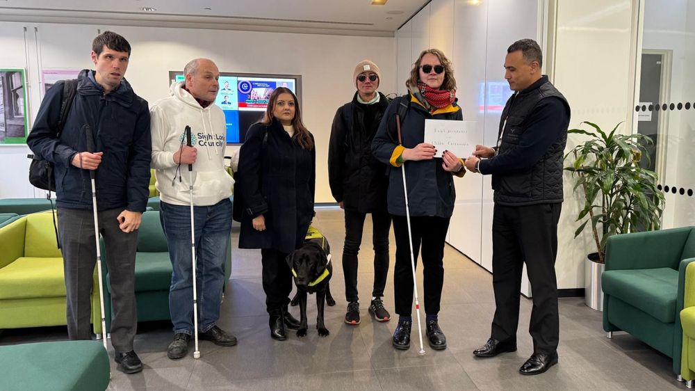 Andy Law, Policy and Campaigns Manager at TPT, Martha Foulds, Policy and Campaigns Coordinator at TPT, Paul Hsu, North London SLC, Bhavini Makwana, Policy and Campaigns Manager at TPT, Liam O'Carroll, Interim Engagement Manager for London Sight Loss Councils', and Shalini O'Kane, SW London SLC member are standing indoors in the Department for Transport with a card addressed to Heidi Alexander and Lilian Greenwood. They are handing it to a member of the department.