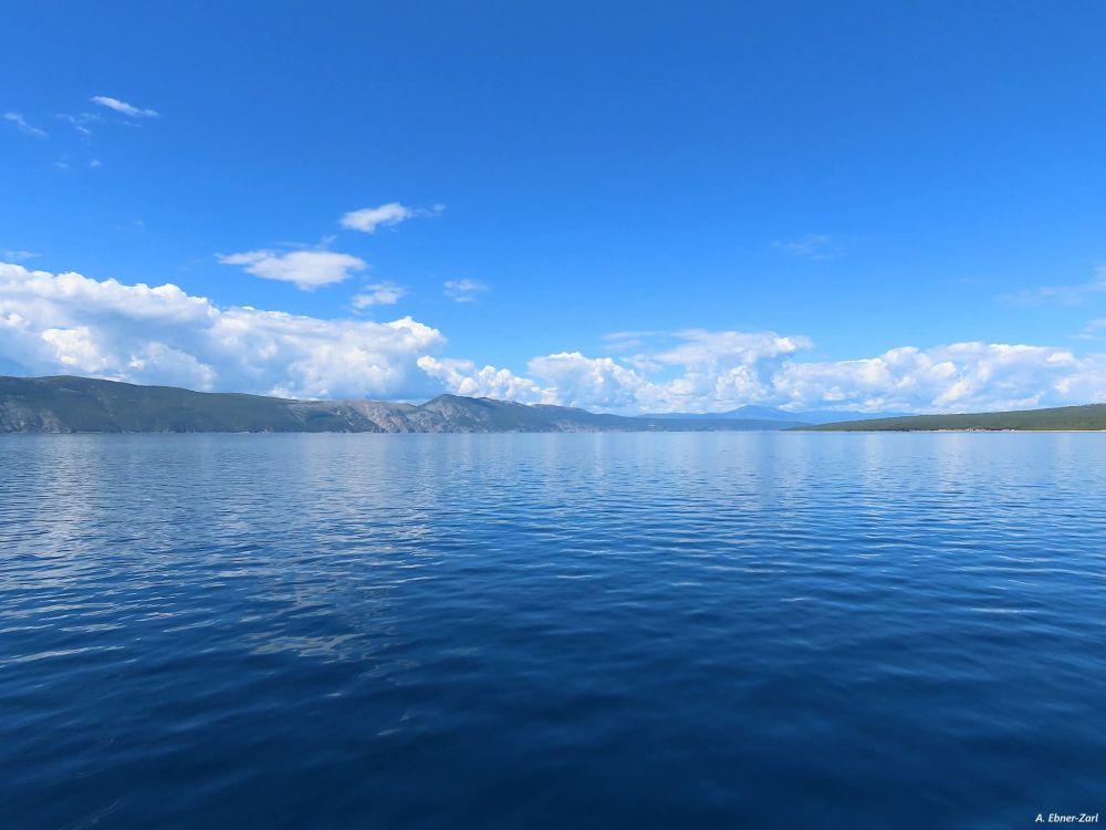 Photo of a deep blue and very smooth sea surface, lightly reflecting the white clouds and the bodies of land on the horizon. 
