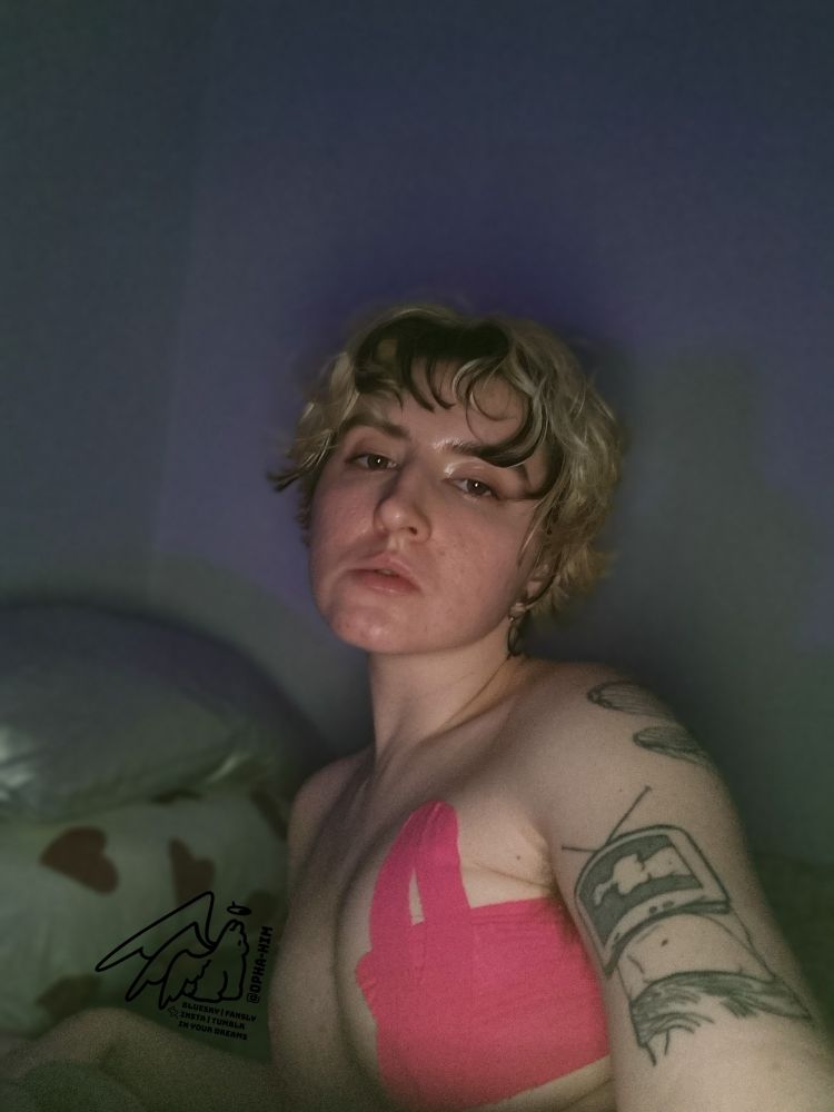 A selfie of Raz inside a dark room with a purple tint to the shadows on the wall behind him. You can see his tattoos on the arm holding the phone, and pink kinesiology tape binding his naked chest. In the background are two pillows, of which one has pink hearts on it.