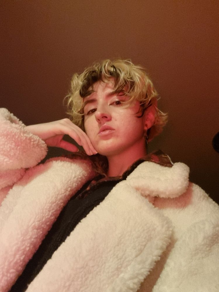 A low angle selfie of Raz, a trans man with a pale complexion and bleached short, wavy hair with a dark under layer wearing a puffy white coat, a black sweater vest with a dress shirt with a red snake pattern underneath. He's doing another pose, this time with his fingers backsides resting on against face.