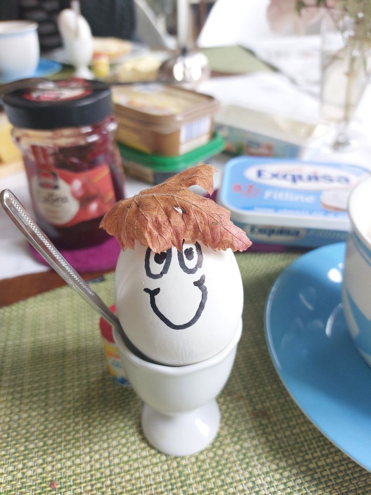 Ein Ei im Becher auf einem Frühstückstisch mit welkem Herbstblatt auf dem "Kopf" und aufgemaltem freundlichen Gesicht.