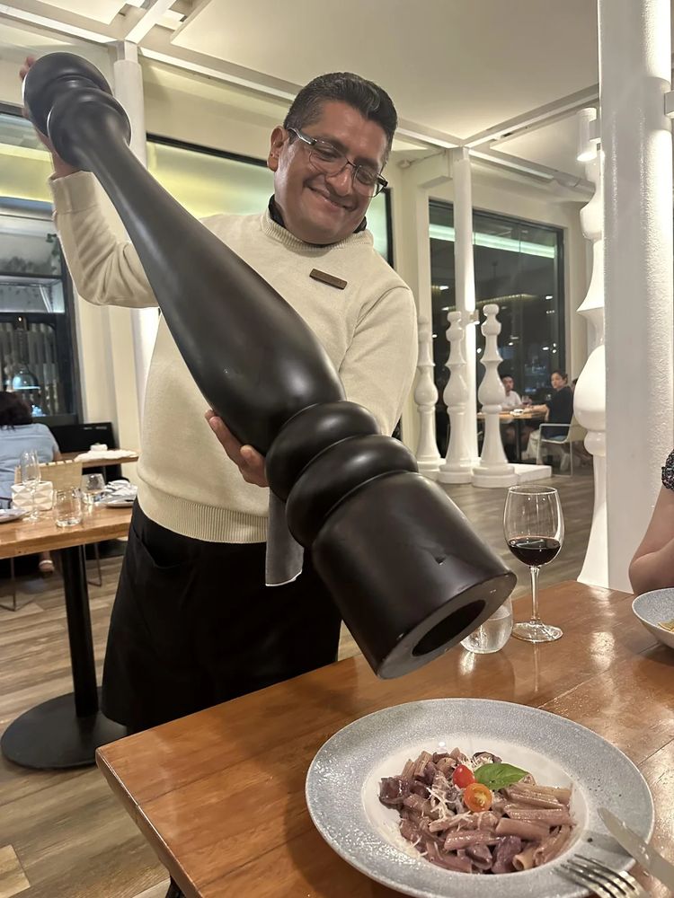 An image of a man using a frankly colossal pepper grinder on a tiny plate of food, it can't be overstated how large the grinder is, it's just bloody enormous. Like a meter or something hot damn