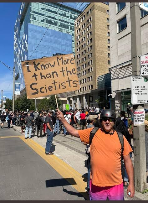 A douchebag cooker antivaxxer at a rally holds a sign saying ‘I know more than the scietists’
