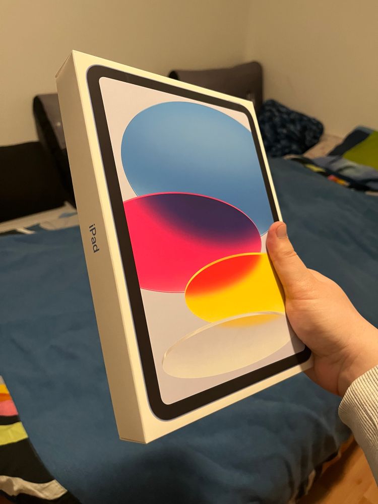 Emma holding up an unopened box of iPad. It is the basic model with the A16 chip, and is in blue color. In the background, her bed is visible.