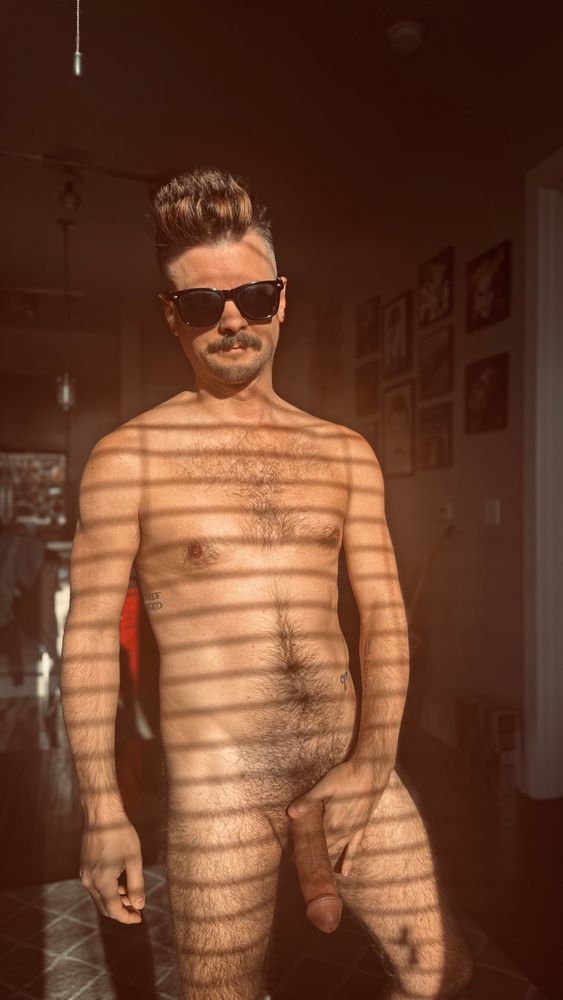 A shot of me standing naked in front of my window with the bright light pouring in on my hairy body. I’m wearing a pair of sunglasses and holding my fat hard cock.