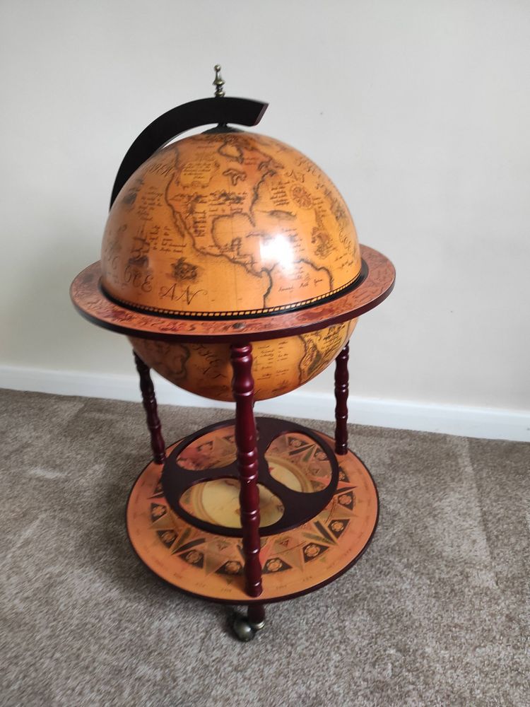 A wooden painted wooden globe in sepia and brown tones on three castered legs with a space to store bottles between the legs