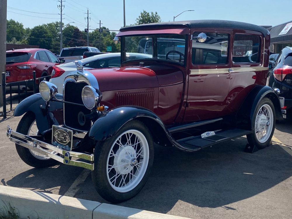 Front 3/4 view of a maroon 1929 Model A Ford two door sedan with black fenders and white wire wheels. It is in the inventory of a used car lot.