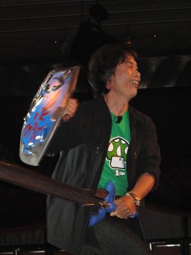 Shigeru Miyamoto on stage at E3 in 2005, wearing a 1UP t-shirt and carrying a Zelda sword and shield.