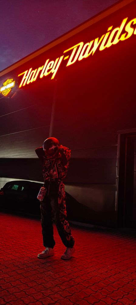 aforementioned tgirl now also wearing a motorcycle helmet standing in front of a huge harley-davidson sign, the source of the orange light in all other photos