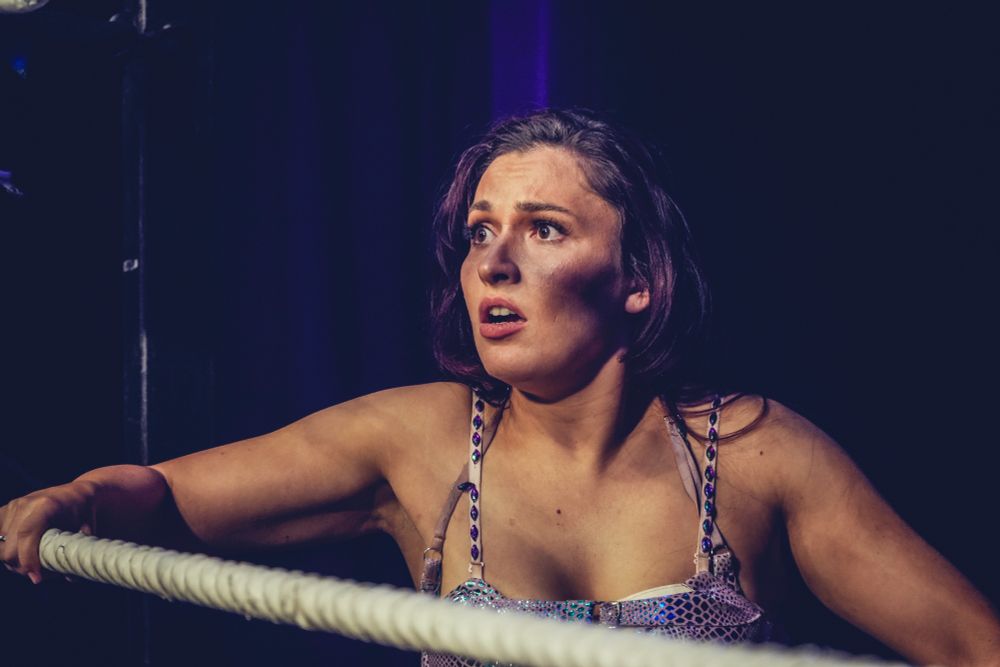 A female wrestler stands outside the ring, holding the bottom rope and looking in