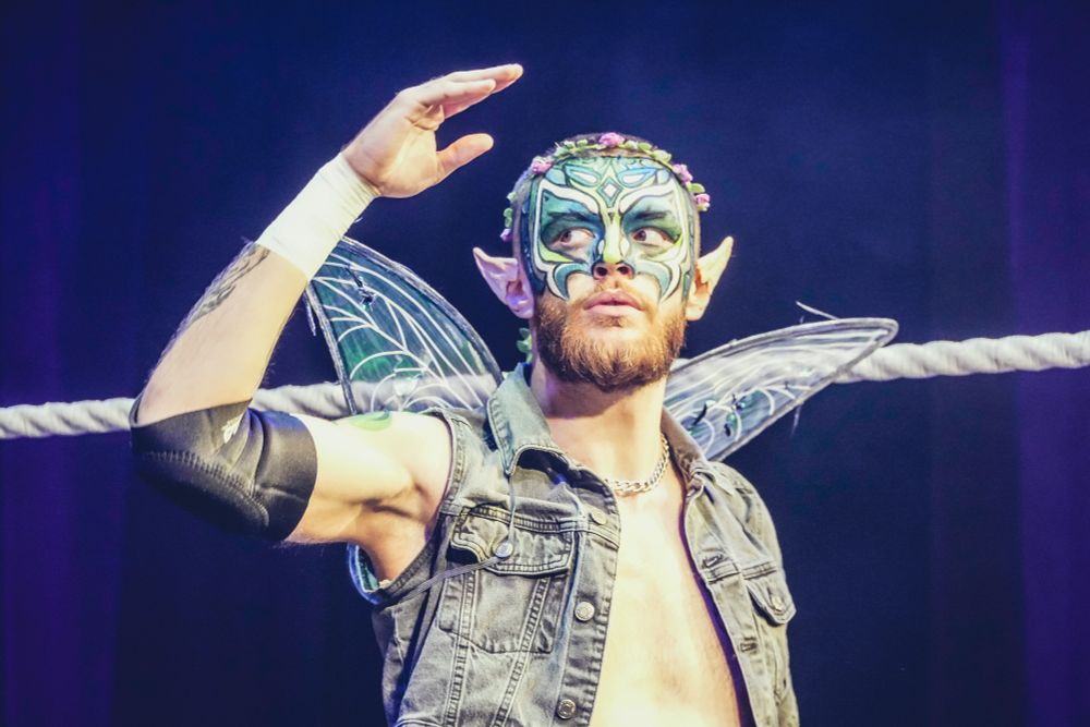 A wrestler with fairy wings and blue and gold makeup raises his right hand in salute