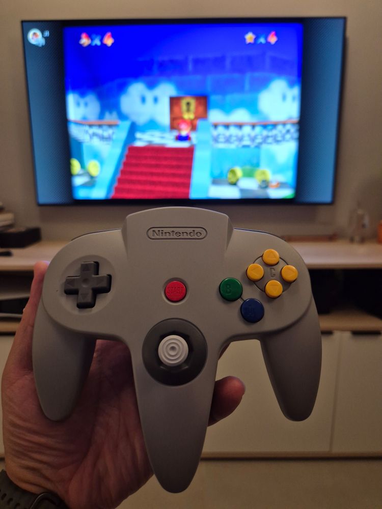 Controle do Nintendo 64 na minha mão em primeiro plano, no fundo Mario 64 rodando na TV