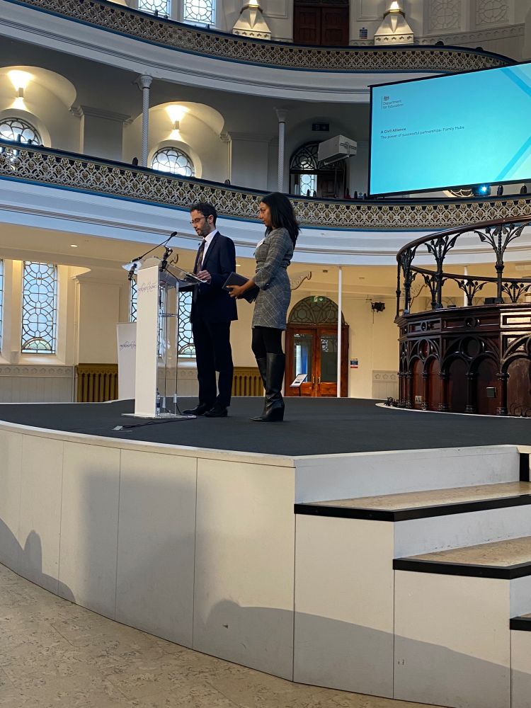 Two people speak on stage, in front of a Department for Education screen