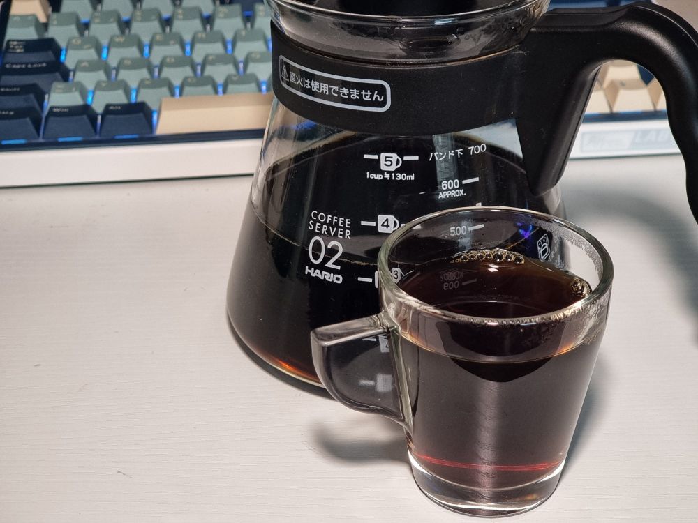A little glas cup filled with filtered coffee in front of a Hario V60 02 coffee server. The coffee has a slight reddish brown shimmer. In the background you can see my light blue mechanical keyboard on my desk.