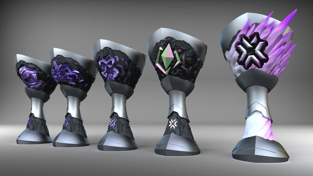 A line of 5 esports trophies.  The cup shape is a satin silver with black stone accents.  The top of the cup houses an element from the game Valorant.  From left to right: a purple oni mouth mask, purple butterflies with electric bolts, a purple repeating hexagonal pattern, a yellow/blue/pink color shift diamond and electric jolts, and a white to pink spiky crystal. 