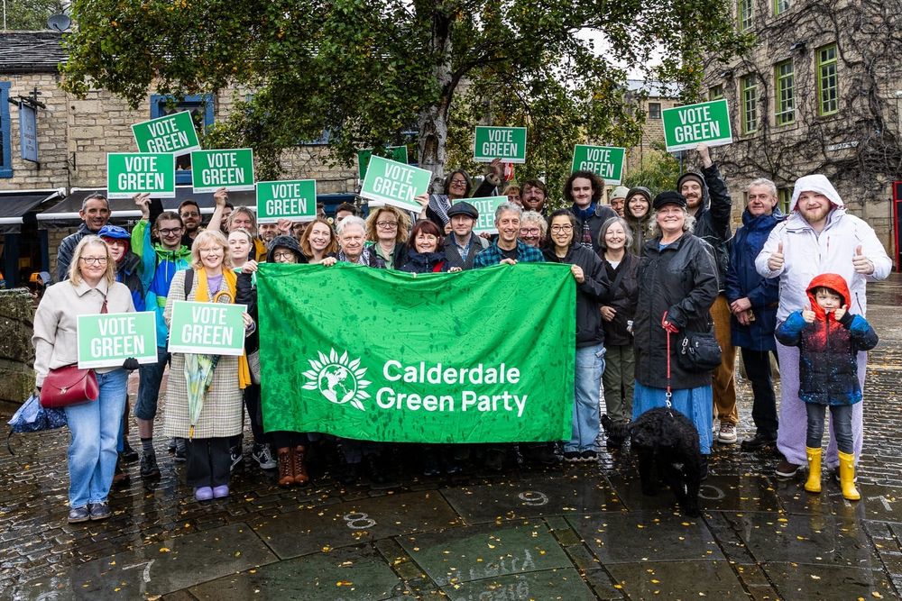 A group shot of Calderdale Green Party members holding signs saying "vote Green" on a rainy day.