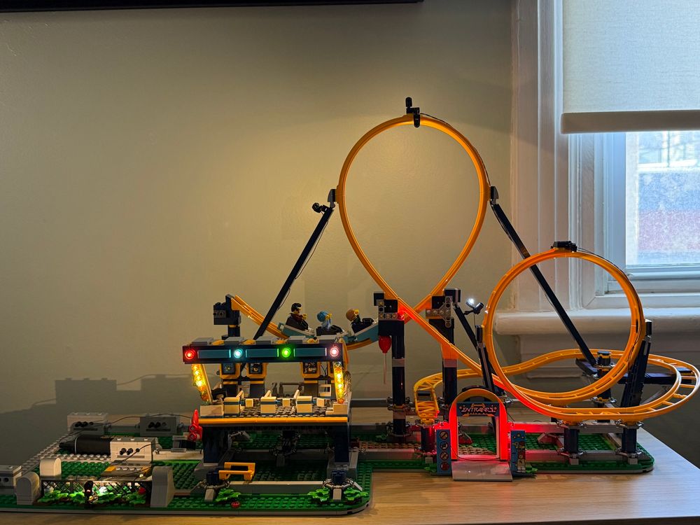 The right side of the LEGO amusement park is now fully illuminated, setting the stage for maximum thrills—or impending disaster. The roller coaster station glows with colorful lights, casting a warm and inviting ambiance, while the towering yellow loops gleam ominously in the background. A roller coaster car filled with nervous minifigures sits at the top of the first drop, their expressions ranging from excitement to deep regret. Meanwhile, red warning lights flash near the entrance, possibly signaling that something is very wrong. Could this be the work of the disgruntled saboteur from earlier? The scene is cinematic, dramatic, and full of anticipation—either this will be the best ride of these minifigures’ lives, or an unforgettable disaster in the making.