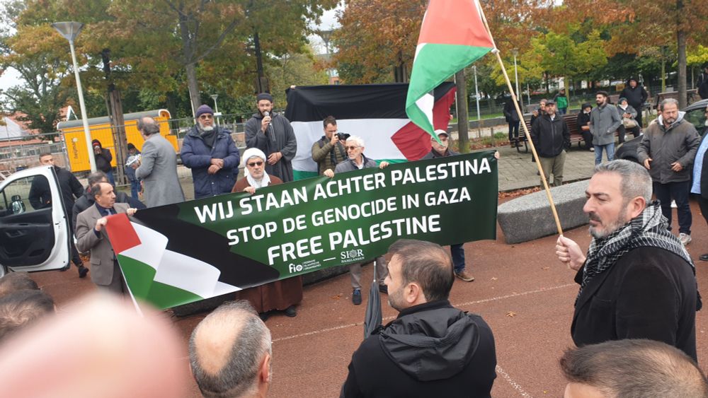 Foto met verscheidene mensen bijeen in demonstratie, op het wijkpark Transvaal, o.a. een Imam die aan het woord is, een man met Palestijnse vlag, een grote Palestijnse vlag en een spandoek met daarop 'Wij staan achter Palestina. Stop de Genocide in Gaza. Free Palestine.' En daaronder organisatoren: FIO Federatie Islamitische Organisties - SIOR Haaglanden