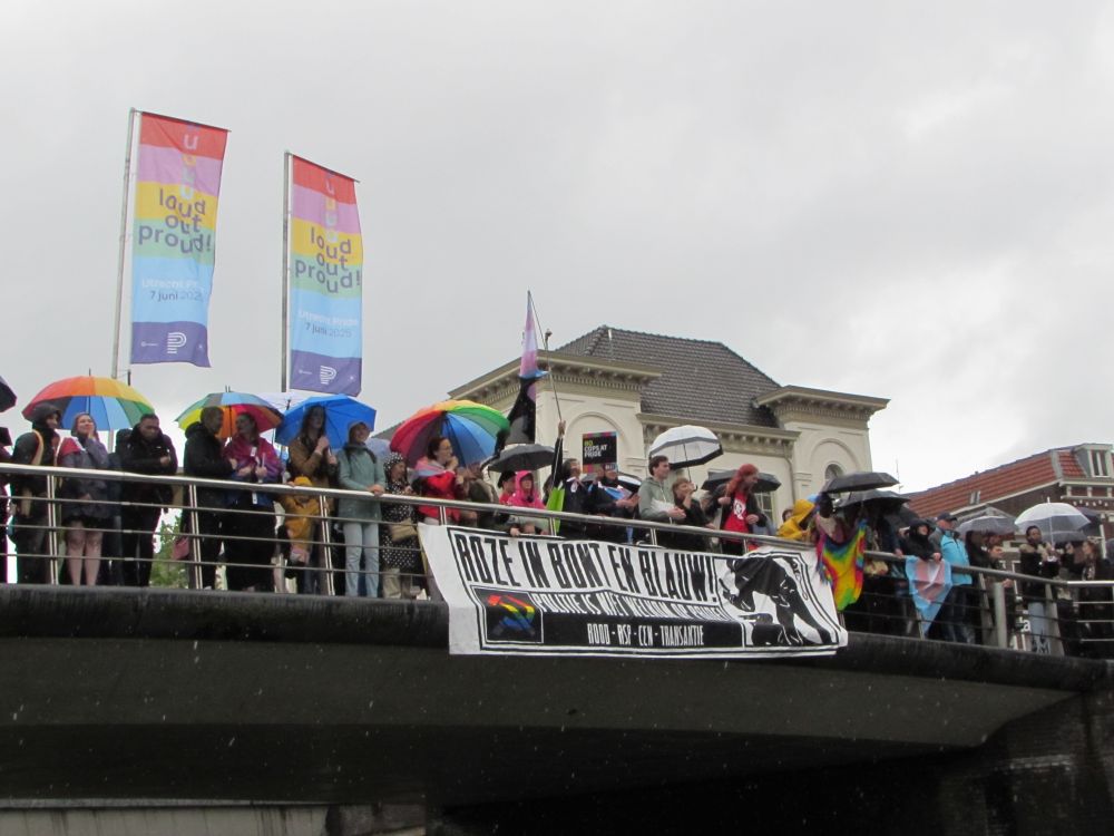 A banner dropped from a bridge. The banner says "Roze in bont en blauw! Politie is niet welkom op pride. ROOD - RSP - CCN - TransAktie"