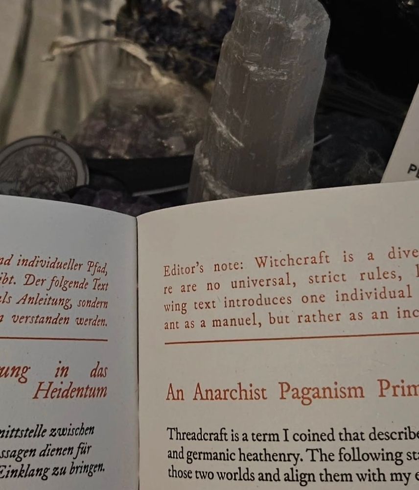 A photo of the upper center of two pages in the zine — a few words in an orange font are revealed, most notably a part of an editors note and the subheading in english: "An Anarchist Pagan Primer". Behind the opened Zine in the background are a few purple and white crystals, a bundle of lavender, and a silver charm blurred because of the focus on the pages.