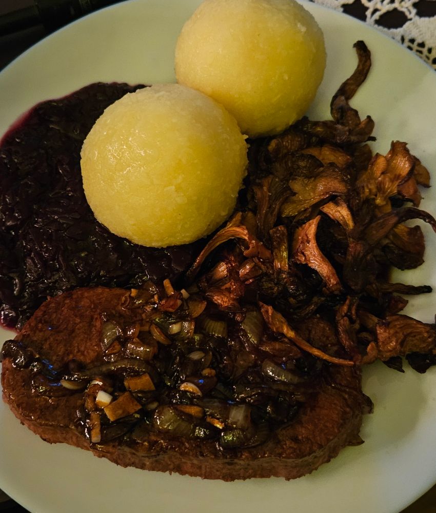 Foto eines Tellers mit Klößen, kross gebratenen Pfifferlingen, dunklem Rotkraut, und einem dunklen Veganen Steak getoppt mit ein paar dunklen Zwiebeln und Knoblauch. Es sieht alles sehr saftig und gesättigt aus und die Klöße stoßen golden hervor.