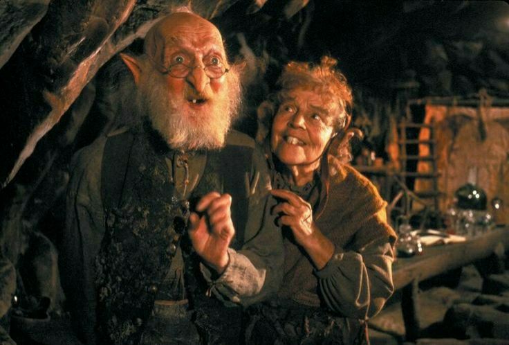 Engywook and Urgl from The Neverending Story movie part 1.
two small goblin-like characters. Engywook has only one tooth and Urgl ha bunny-teeth. They both have messy hair and pointy ears, while Engywook only has a few ears on his head. Engywook wears thin glasses on his nose. They stand in their little hut with warm light from a fire and a table.