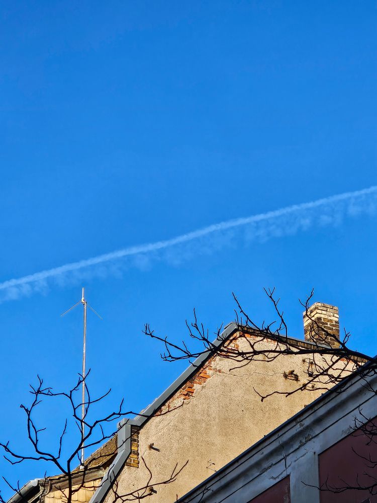Photo of a roof from the side (rooftop not visible). The blue sky above is ornamented with a faded airplane stripe. The side of the roof looks old, the plaster is falling of in some areas revealing red bricks. A few leafless branches and a stalk-like wind measuring device adorn the image of the roof.