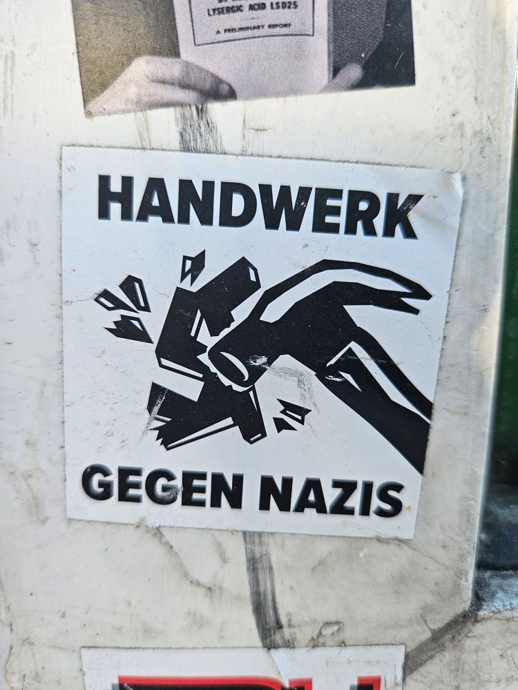 Photo of a black an white sticker, stuck on a dirty white wall, that reads "Handwerk gegen Nazis" (craftmanship against Nazis) with a hammer smashing a Hakenkreuz. 